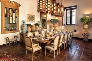 Un comedor con una larga mesa de madera dispuesta para una comida, rodeada de sillas, con un espejo y jarrones decorativos en las paredes.