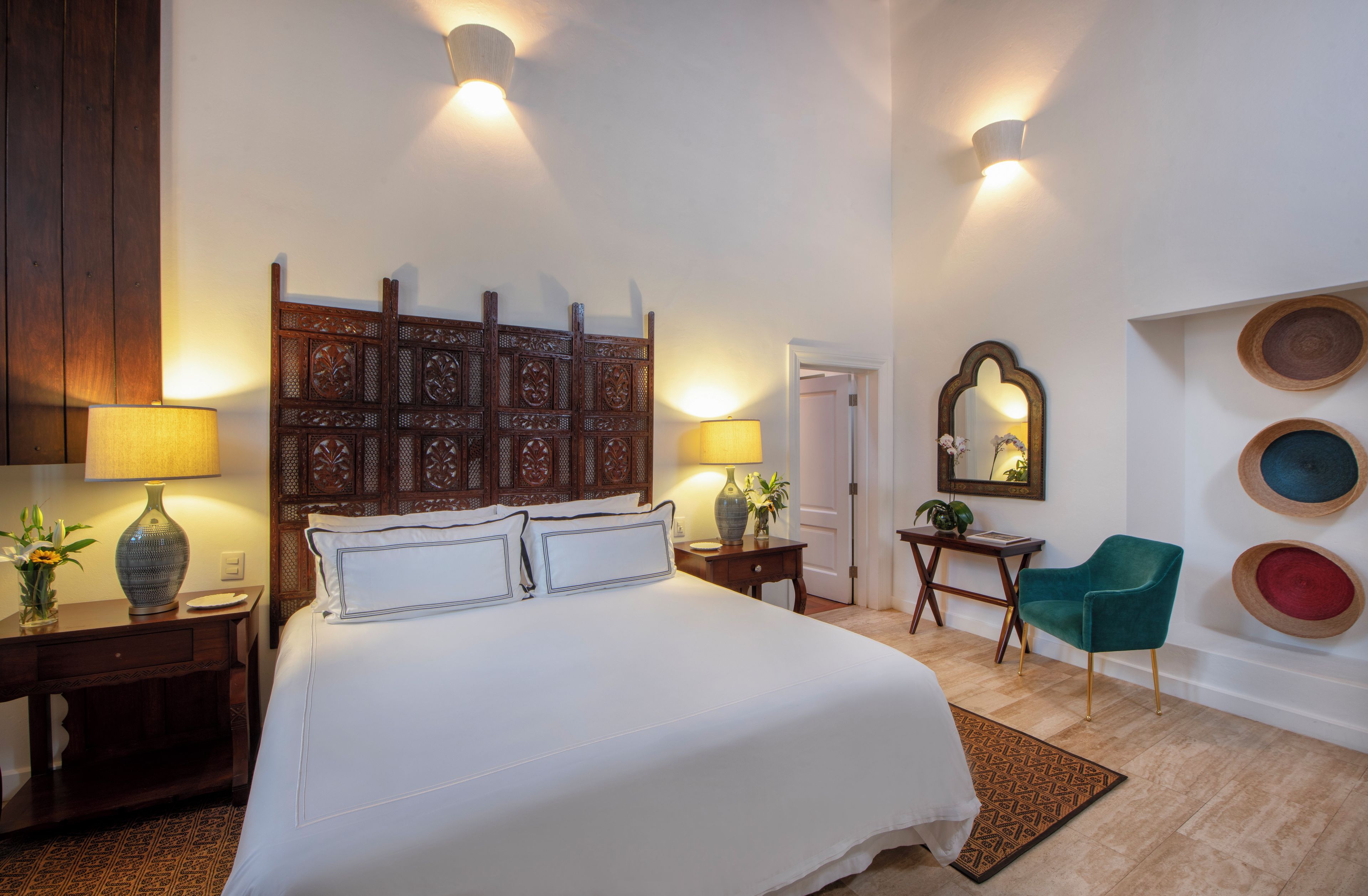 Une chambre d’hôtel avec un grand lit, deux tables de chevet avec des lampes, une tête de lit en bois, un miroir, une chaise et des éléments décoratifs fixés au mur.