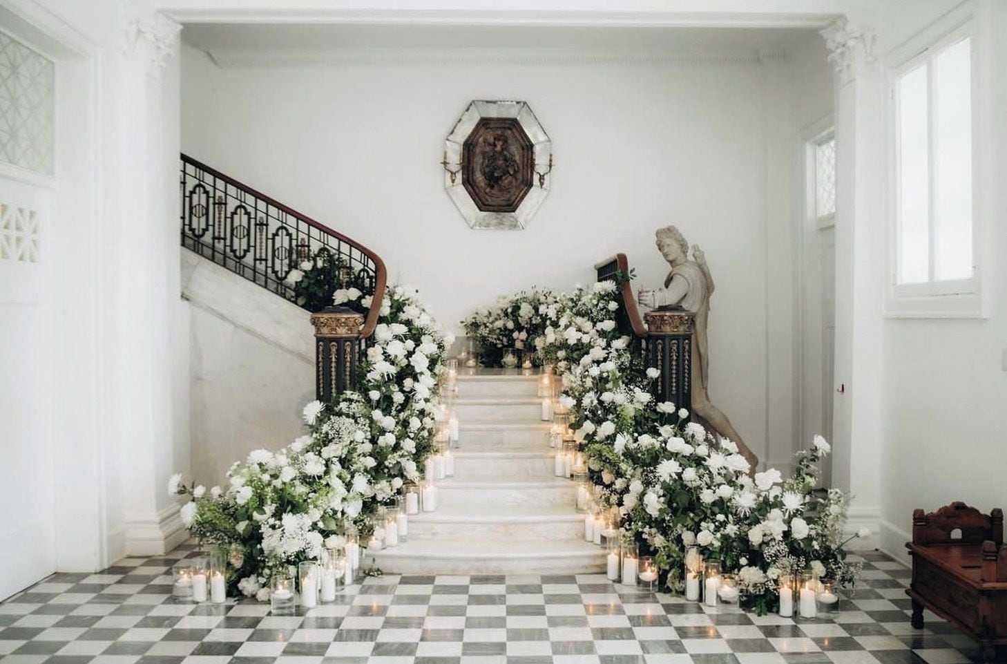 A grand staircase with white floral arrangements and candles, leading to a landing with a bust sculpture.