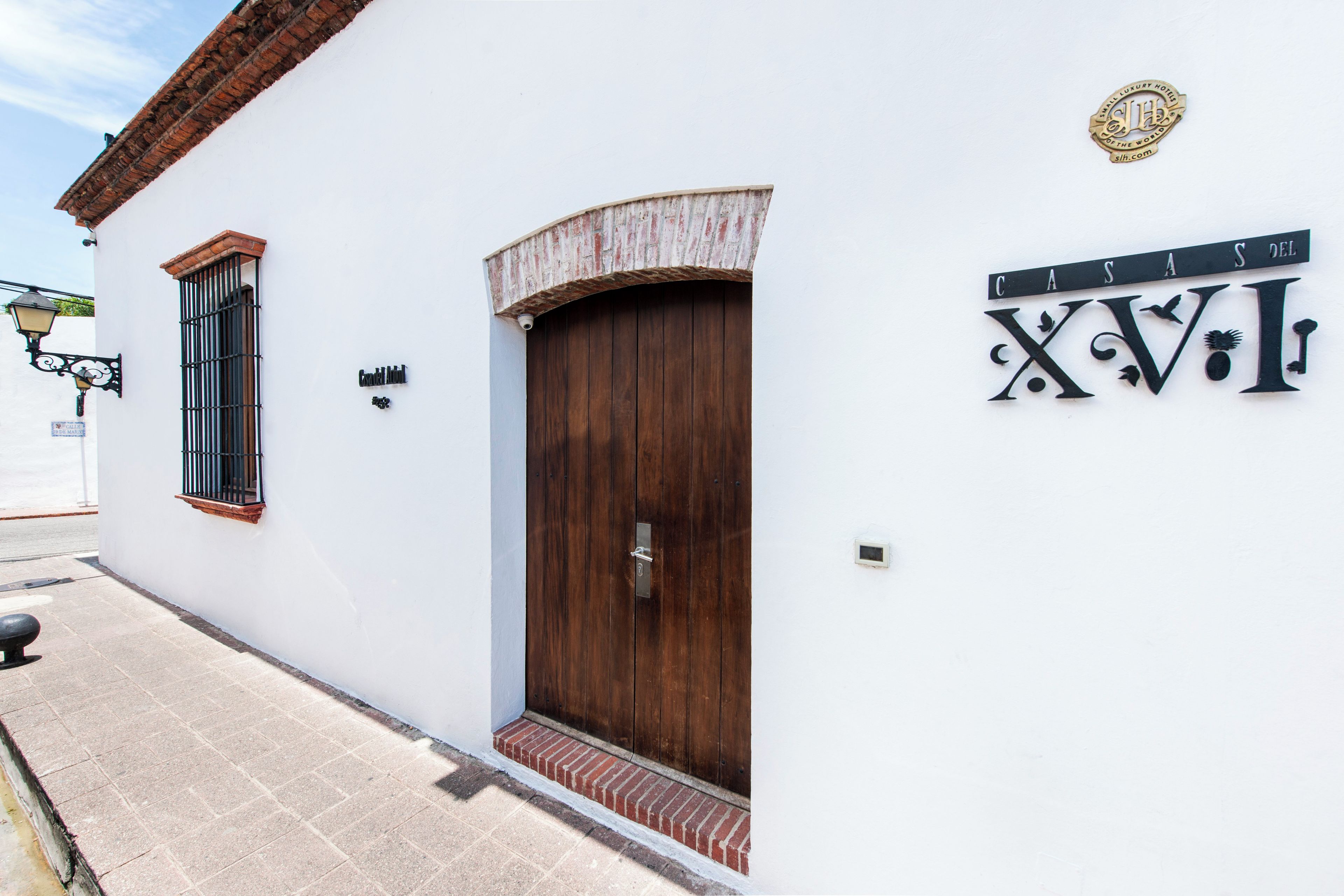 A white building with a wooden door and a metal gate, a small sign above the door, and a decorative metal sign with the letters 'XVII' on the wall.