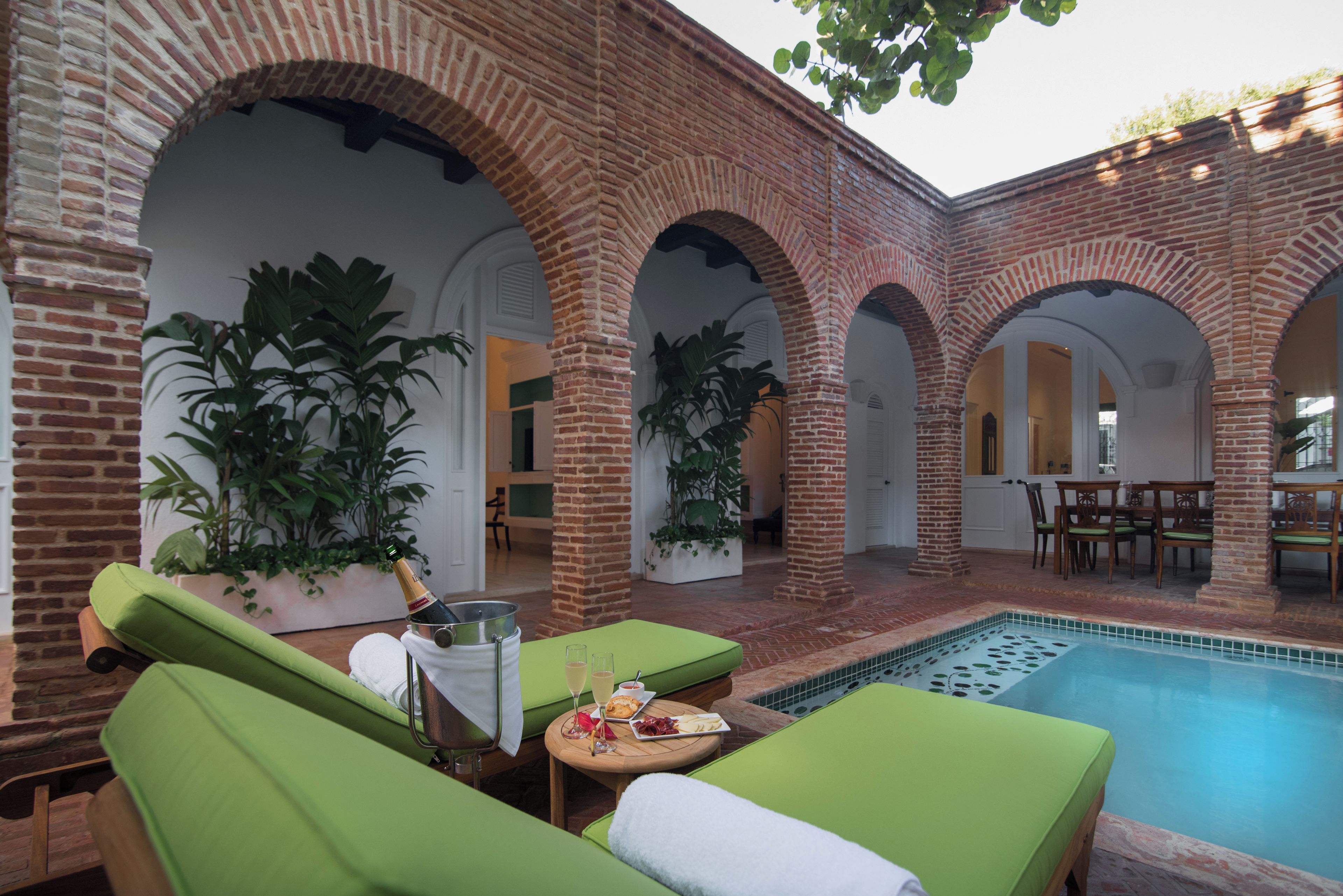 A poolside area with green lounge chairs, a table with a bottle and glasses, and a brick wall with arches.