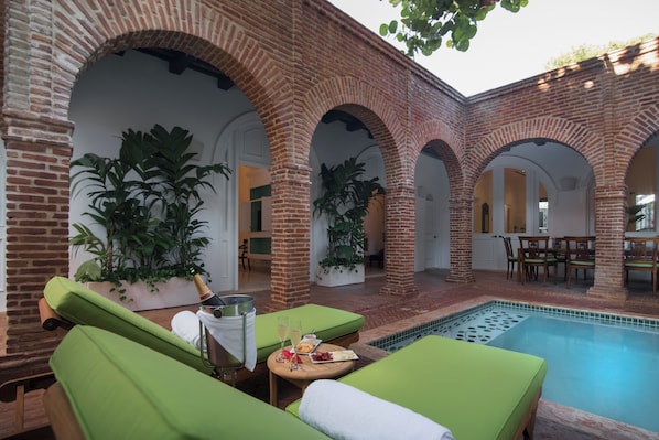 Zona junto a la piscina con sillones reclinables verdes, una mesa con una botella y vasos, y una pared de ladrillo con arcos.
