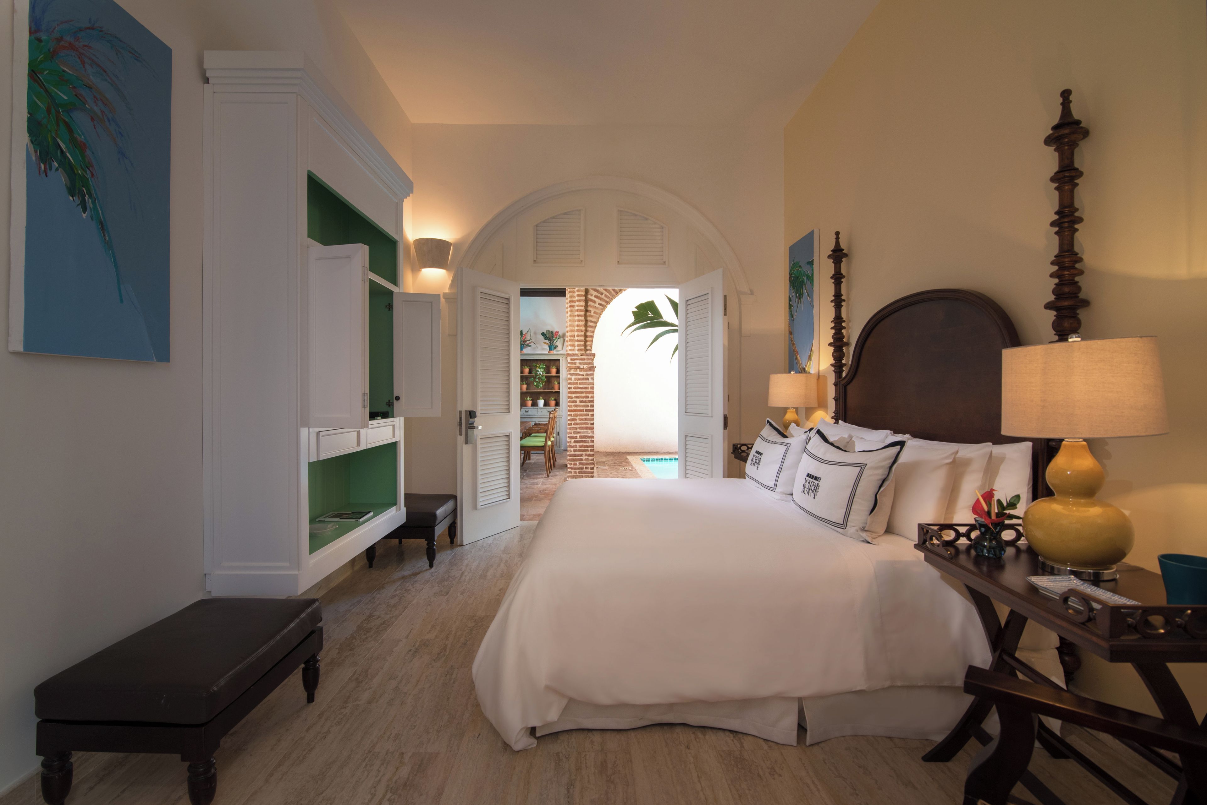 A hotel room with a large bed, a desk with a lamp, a bench, and a view of a pool through an open door.