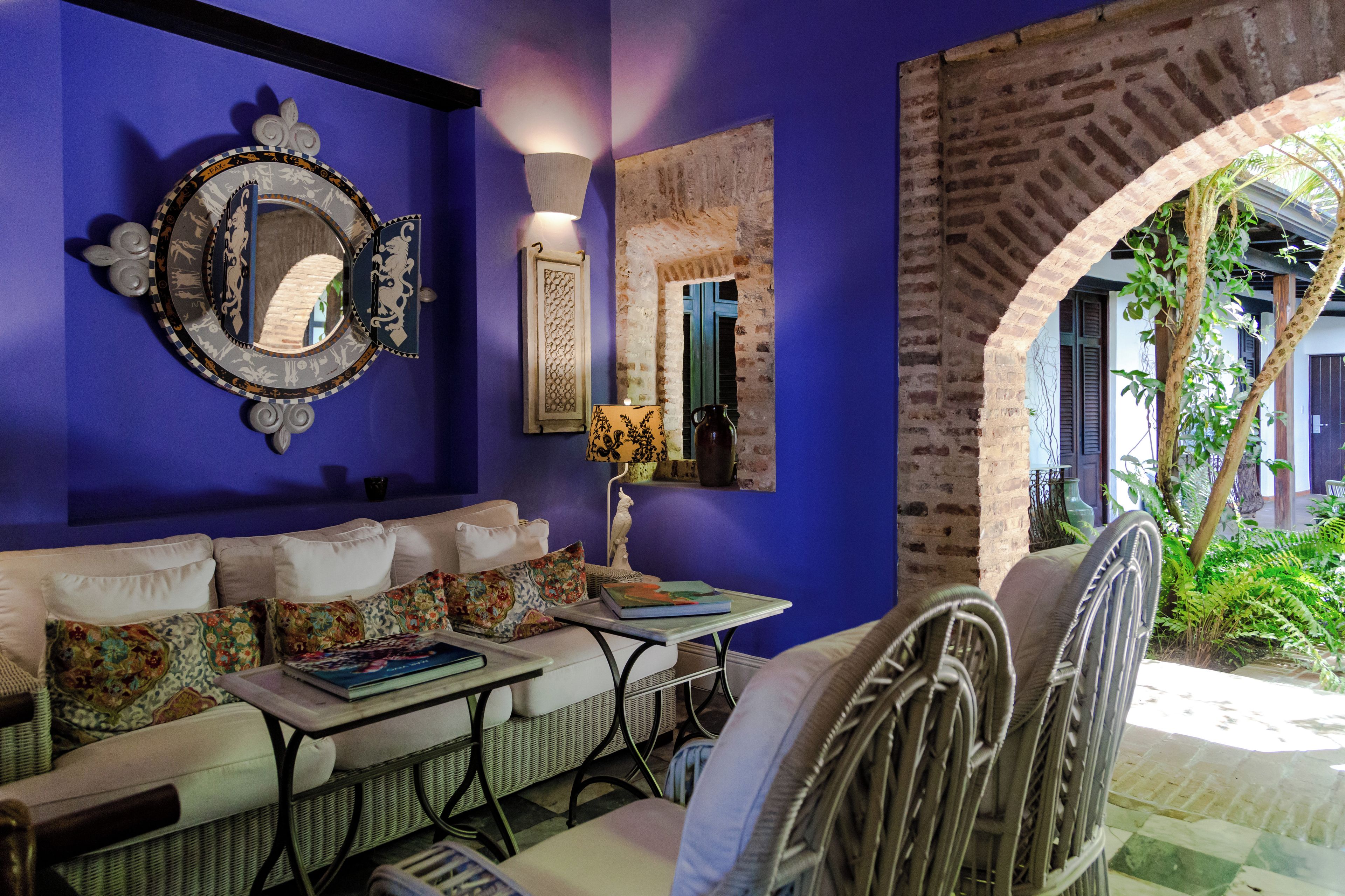 A room with a blue wall, a sofa with patterned cushions, a table with books, a decorative mirror, and a view of greenery through an archway.