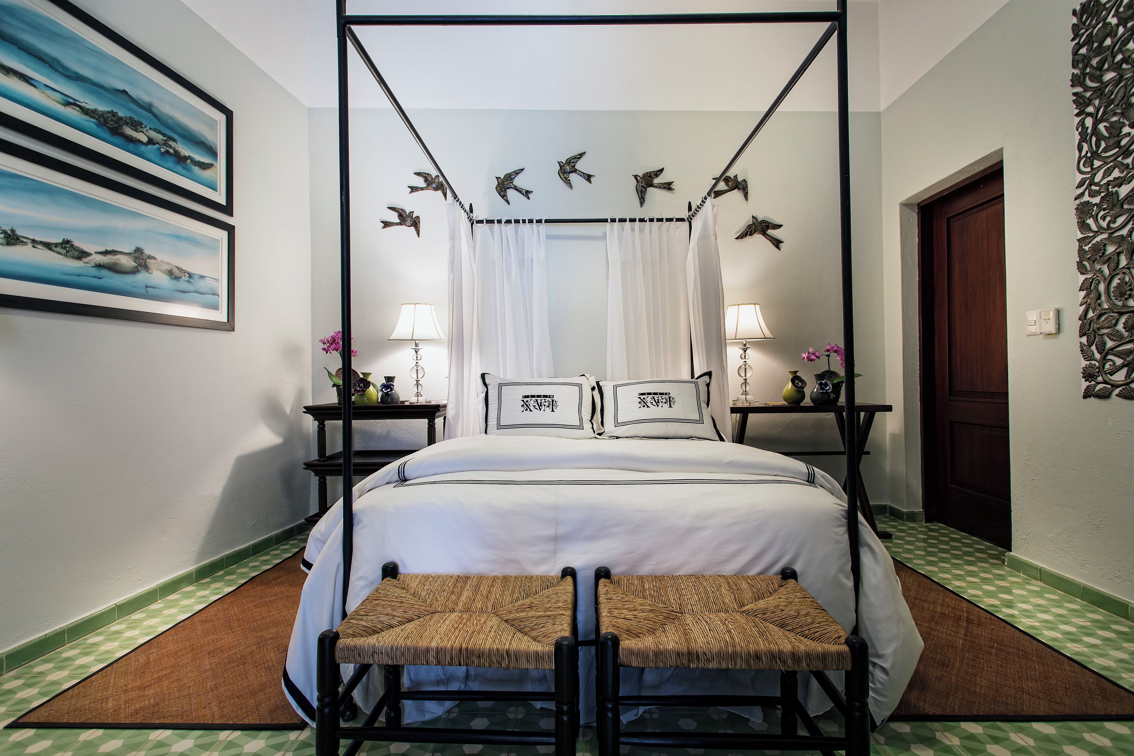 A four-poster bed with a canopy, two bedside tables with lamps, a bench, and artwork on the walls.
