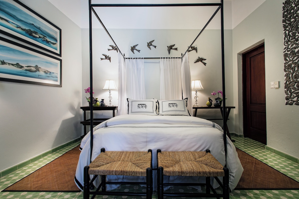 A four-poster bed with a canopy, two bedside tables with lamps, a bench, and artwork on the walls.