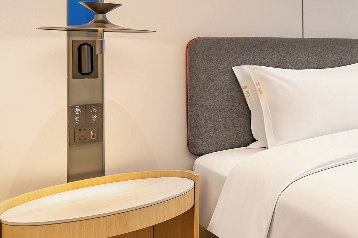 A hotel room with a bed, a bedside table, a lamp, and a control panel.