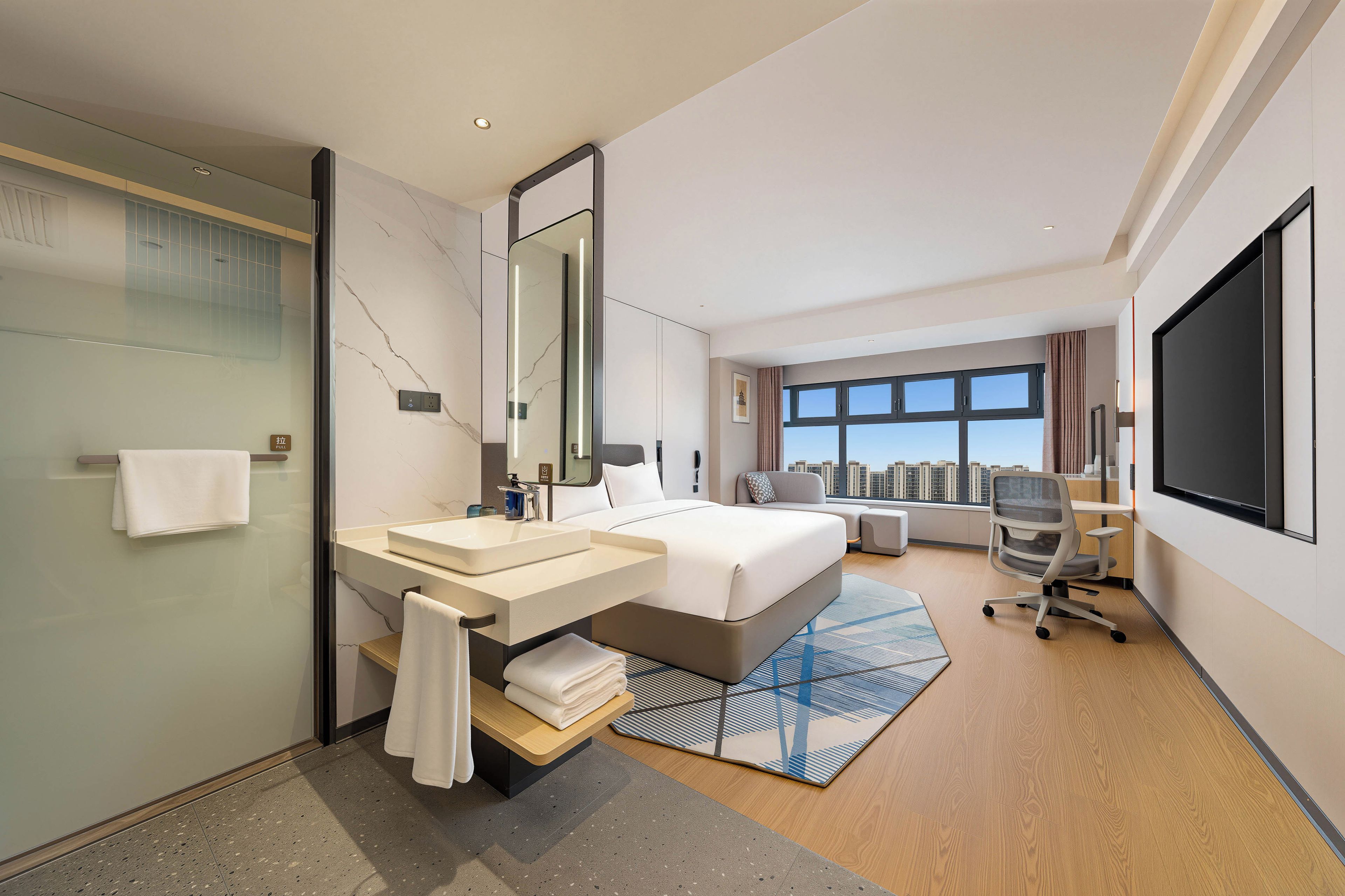 A modern hotel room with a large bed, a desk, a chair, a bathroom with a glass shower, and a city view.