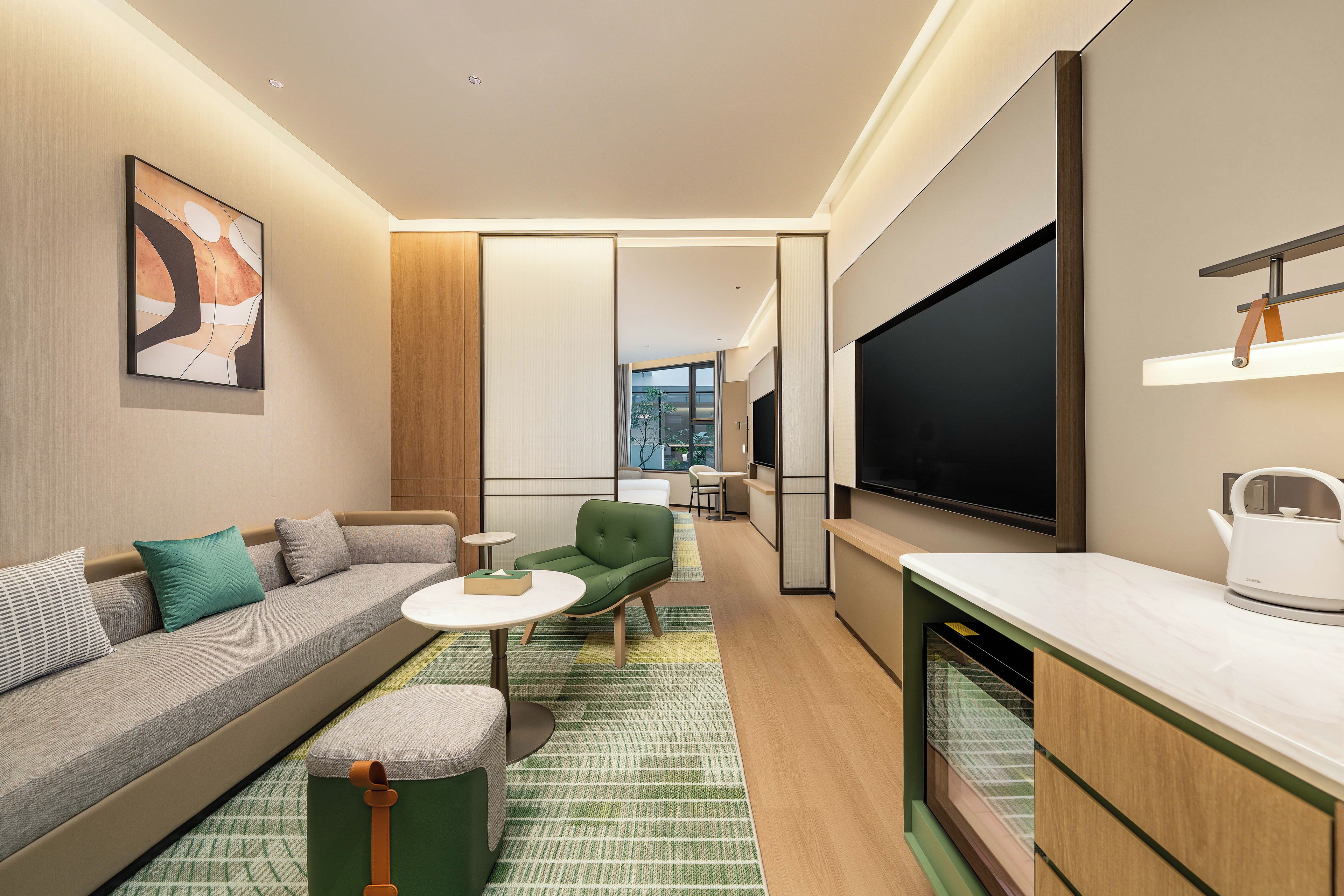 A modern hotel room with a sofa, a small green chair, a table, a flat-screen TV, and a kettle.