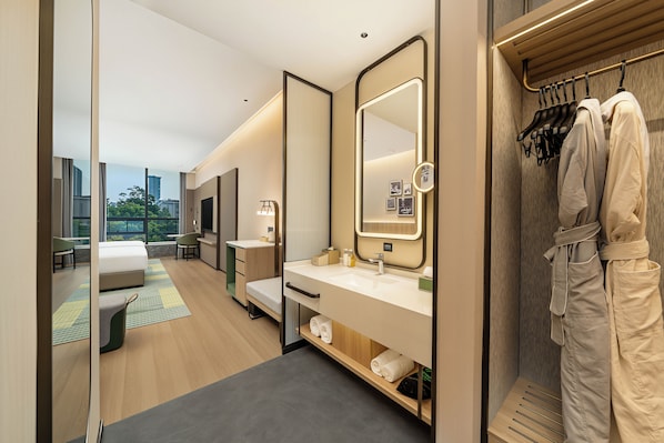 A modern hotel room with a large bed, a sitting area, a vanity with a mirror, and a view of the city.