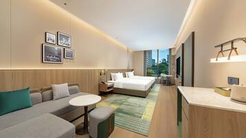 A modern hotel room with a sofa, a bed, a small table, and a desk. There are framed pictures on the wall and a window with a view of buildings.