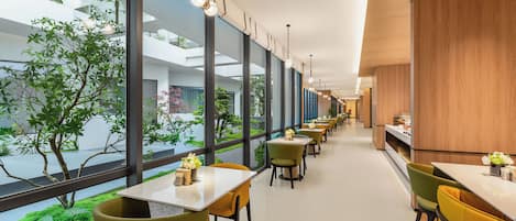 A modern interior with a long corridor lined with tables and chairs, large windows offering views of greenery outside.