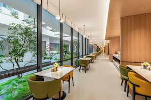 Un intérieur moderne avec un long couloir bordé de tables et de chaises, et de grandes fenêtres donnant sur la verdure environnante.