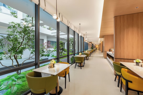 Un intérieur moderne avec un long couloir bordé de tables et de chaises, et de grandes fenêtres donnant sur la verdure environnante.