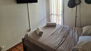 Een slaapkamer met een bed, een flatscreen televisie aan de muur, een raam met gordijnen en een ventilator bevestigd aan de wand.
