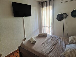 A bedroom with a bed, a flat-screen TV mounted on the wall, a window with curtains, and a wall-mounted fan. - Sea View Apartment with Terraces & Pools (Saint-Mandrier-sur-Mer)