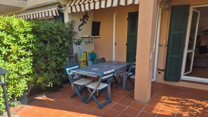 Een terras buiten met een tafel en stoelen, omringd door groen en een betegelde vloer.