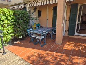 An outdoor patio with a table and chairs, surrounded by greenery and a tiled floor. - Sea View Apartment with Terraces & Pools (Saint-Mandrier-sur-Mer)