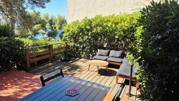 Een terras buiten met een houten tafel, een zithoek met kussens en een houten terras.