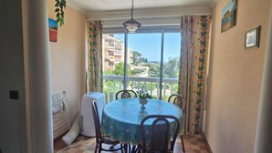 Un coin repas avec une table ronde, des chaises, un ventilateur de plafond et une vue sur les bâtiments à travers une fenêtre ornée de rideaux.