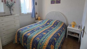 Une chambre à coucher comprenant un lit, une commode, une table de chevet, une lampe, une fenêtre avec des rideaux et un tableau accroché au mur.