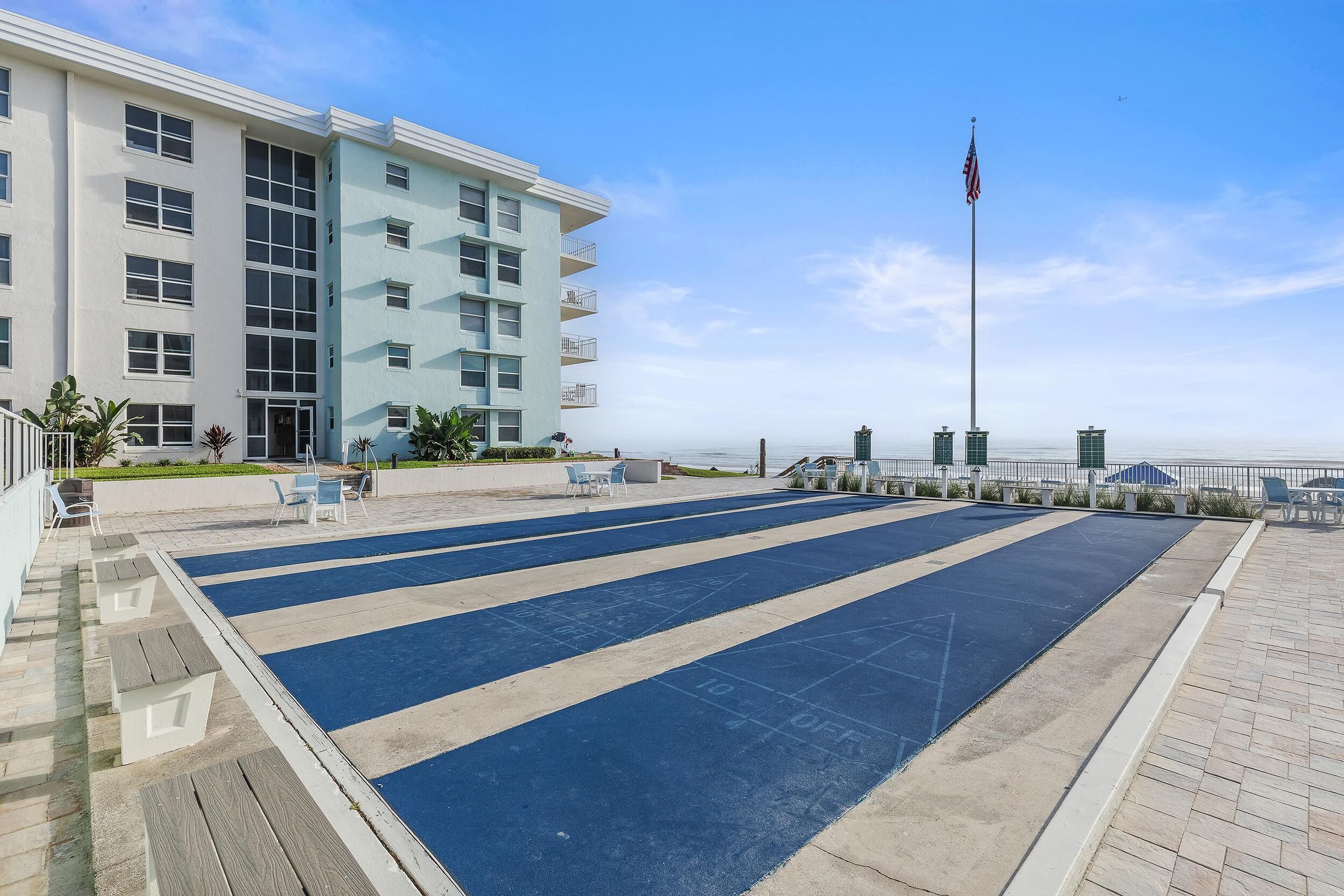 A boardwalk with a blue shuffleboard court, a light blue building with balconies, and an American flag.