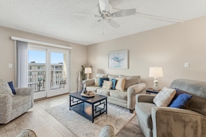 A living room with a ceiling fan, a sofa set, a coffee table, and a view through a sliding glass door. - Sea Coast Gardens II 511 (New Smyrna Beach)