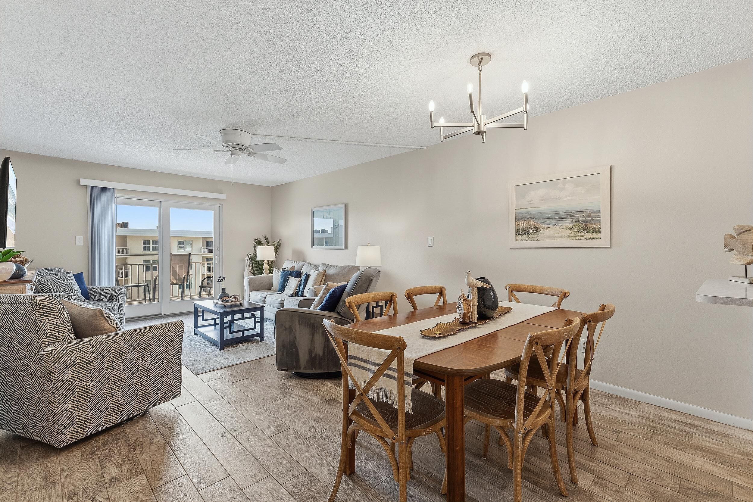 A spacious living room with a dining area, large windows, a ceiling fan, and a painting on the wall.