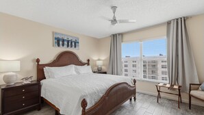 Una habitación de hotel con una cama grande, una mesita de noche, una lámpara, una ventana con cortinas y un ventilador de techo.