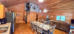 A wooden cabin interior with a dining area, kitchen, and a loft bedroom with a spiral staircase. - Cozy 3-bedroom cabin in gorgeous Little River Canyon with Hot tub (Fort Payne)