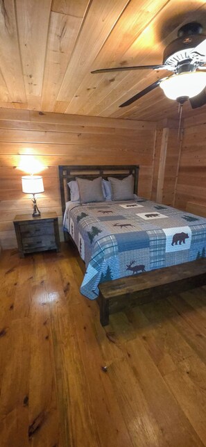 A wooden cabin bedroom with a bed, a nightstand, a lamp, and a ceiling fan. - Cozy 3-bedroom cabin in gorgeous Little River Canyon with Hot tub (Fort Payne)