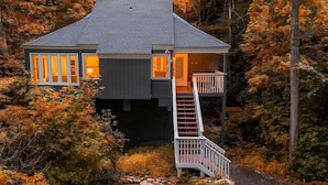 Eine zweistöckige Holzhütte mit einer Holztreppe, die zu einem Balkon führt, umgeben von Herbstlaub.