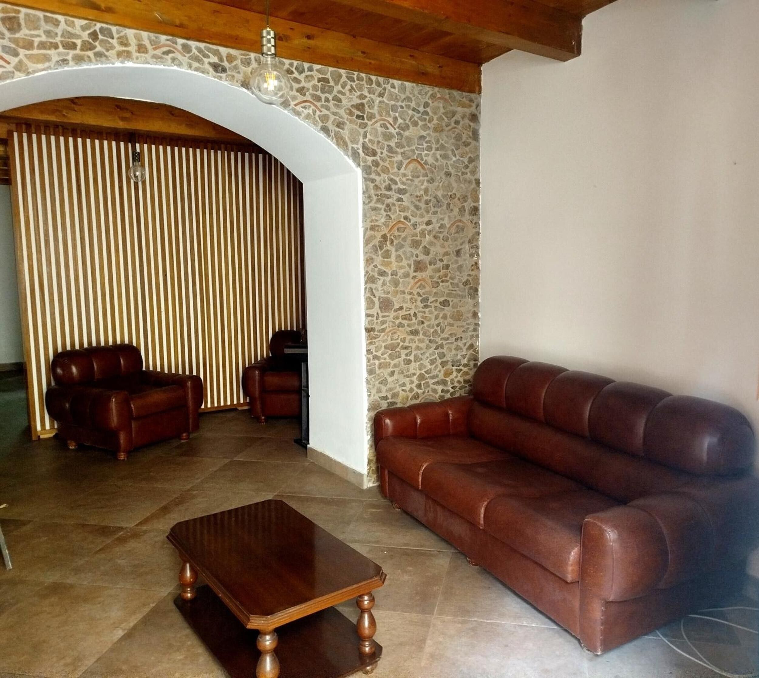 Een woonkamer met een stenen muur, een houten plafond, een bruine leren bank, een houten salontafel en twee leren fauteuils.