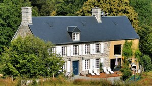 Une maison en pierre à deux étages, avec un toit sombre, des volets blancs et un petit porche agrémenté de chaises.