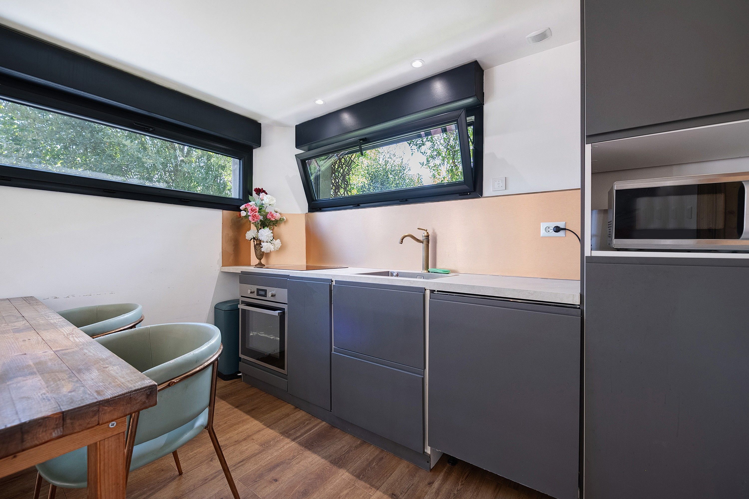Une cuisine compacte équipée d’un four encastré, d’un évier et d’un four à micro-ondes. Une fenêtre se trouve au-dessus de l’évier, et il y a une petite table avec des chaises.