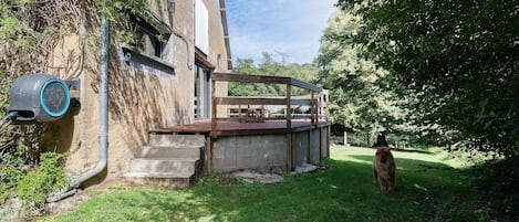 A rustic outdoor area with a concrete staircase leading up to a wooden deck, surrounded by greenery and a building with a window.