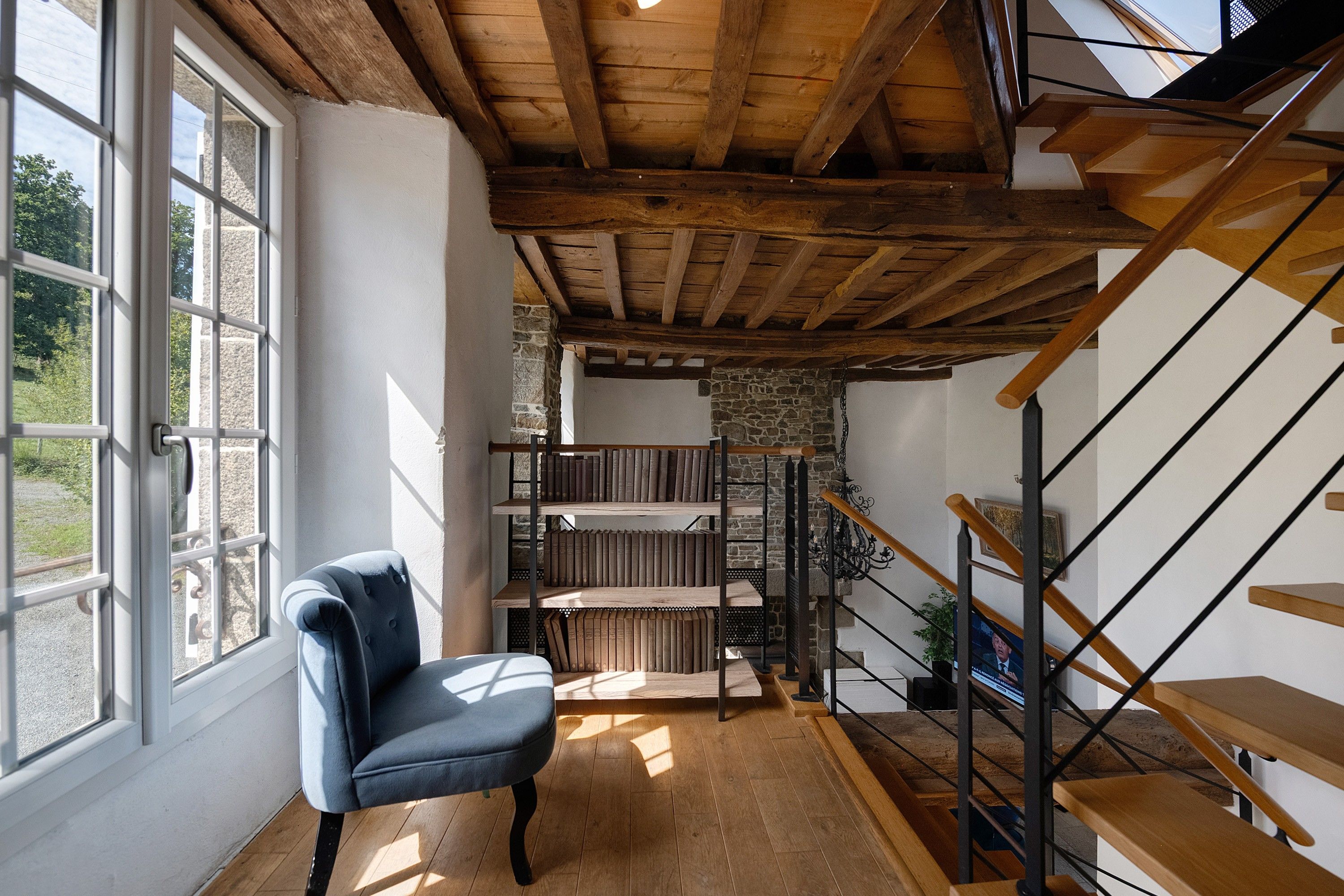 A room with a wooden ceiling, a chair, a staircase, and a bookshelf.