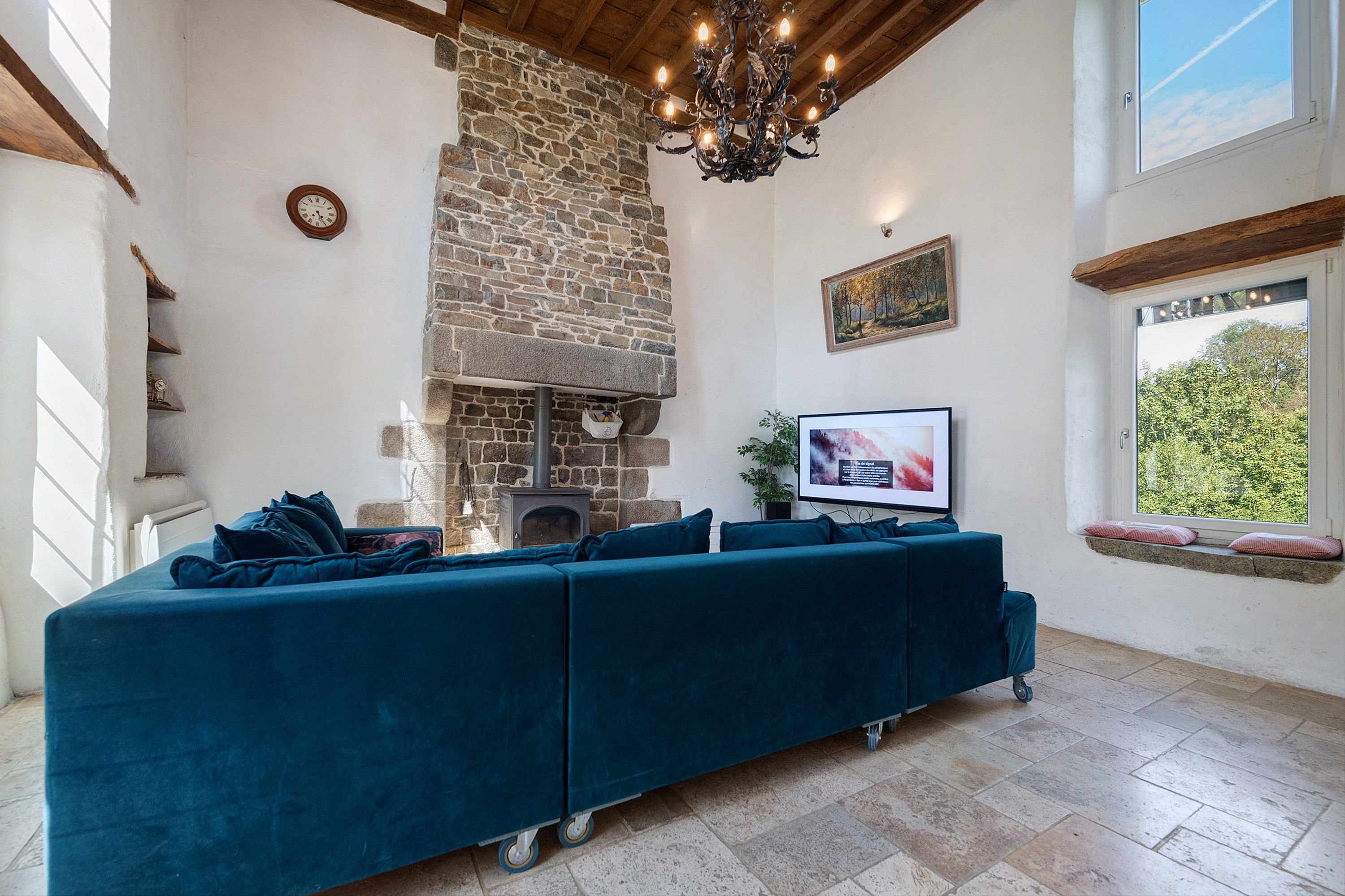 A living room with a stone fireplace, a sofa set, a television, and a window with a view of trees.