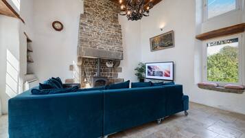 A living room with a stone fireplace, a sofa set, a television, and a window with a view of trees.