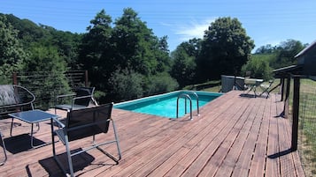 A wooden deck with a swimming pool, surrounded by trees and a stone building.