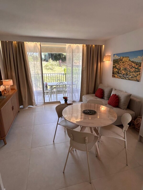 A room with a round table, chairs, a sofa with red cushions, a painting, and a view of greenery through a glass door. - Vrbo Property (Gréoux-les-Bains)
