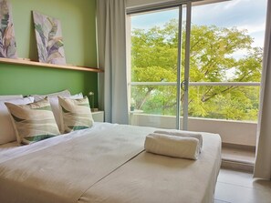 Ein Hotelzimmer mit Bett, Kissen, einem Fenster mit Blick auf Bäume und Kunstwerken an der Wand.