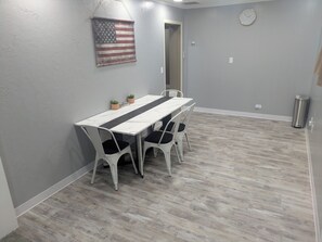Une pièce avec une longue table blanche, quatre chaises blanches, un drapeau américain suspendu au mur et une horloge.