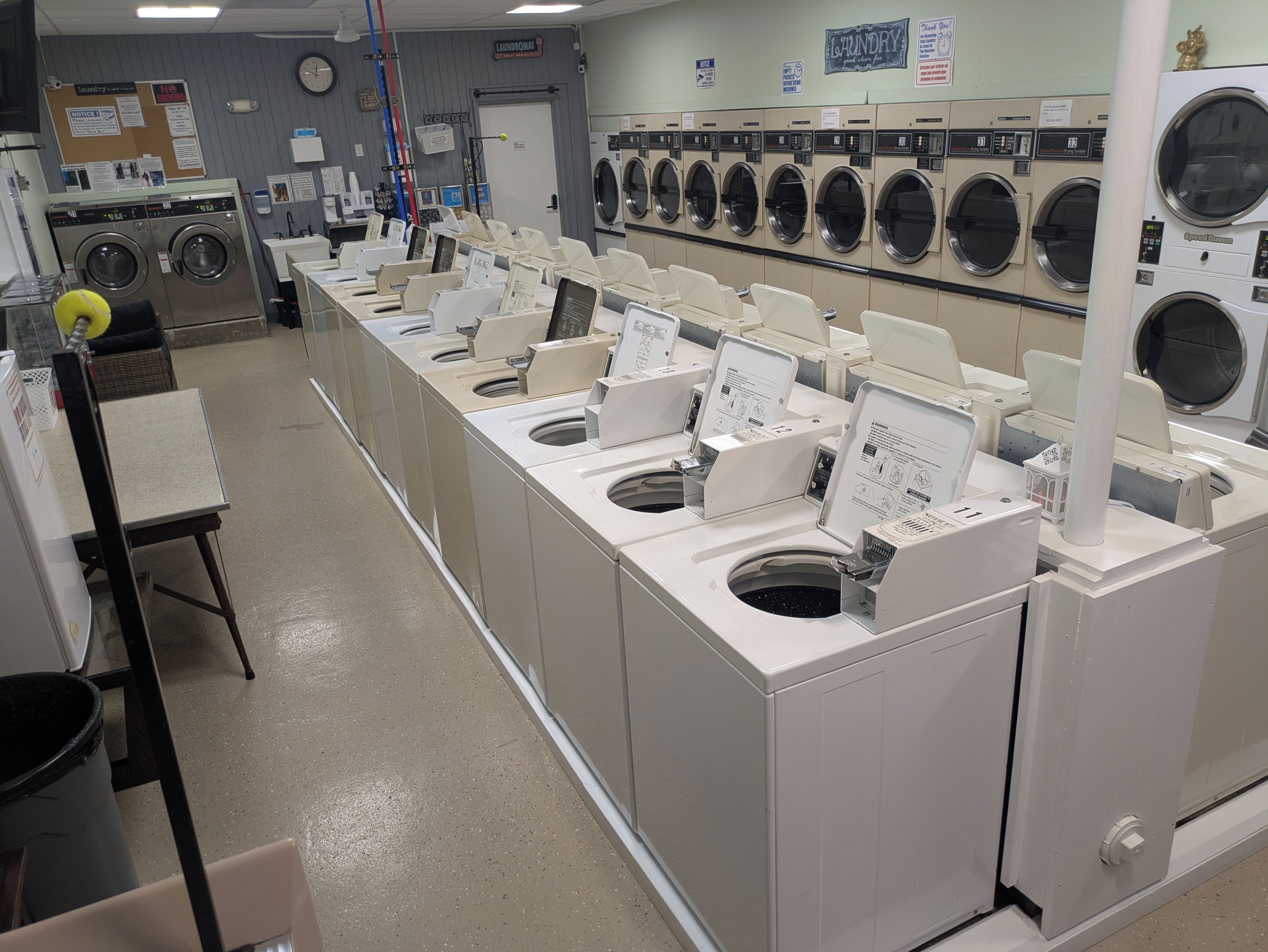 A laundromat with multiple washing machines and dryers, a notice board, and a clock on the wall.