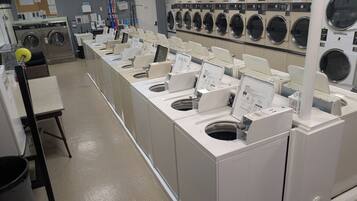 A laundromat with multiple washing machines and dryers, a notice board, and a clock on the wall.