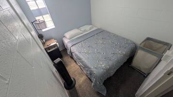 Una habitación pequeña y funcional con una cama individual, una mesita de noche, un cubo de basura y una ventana.
