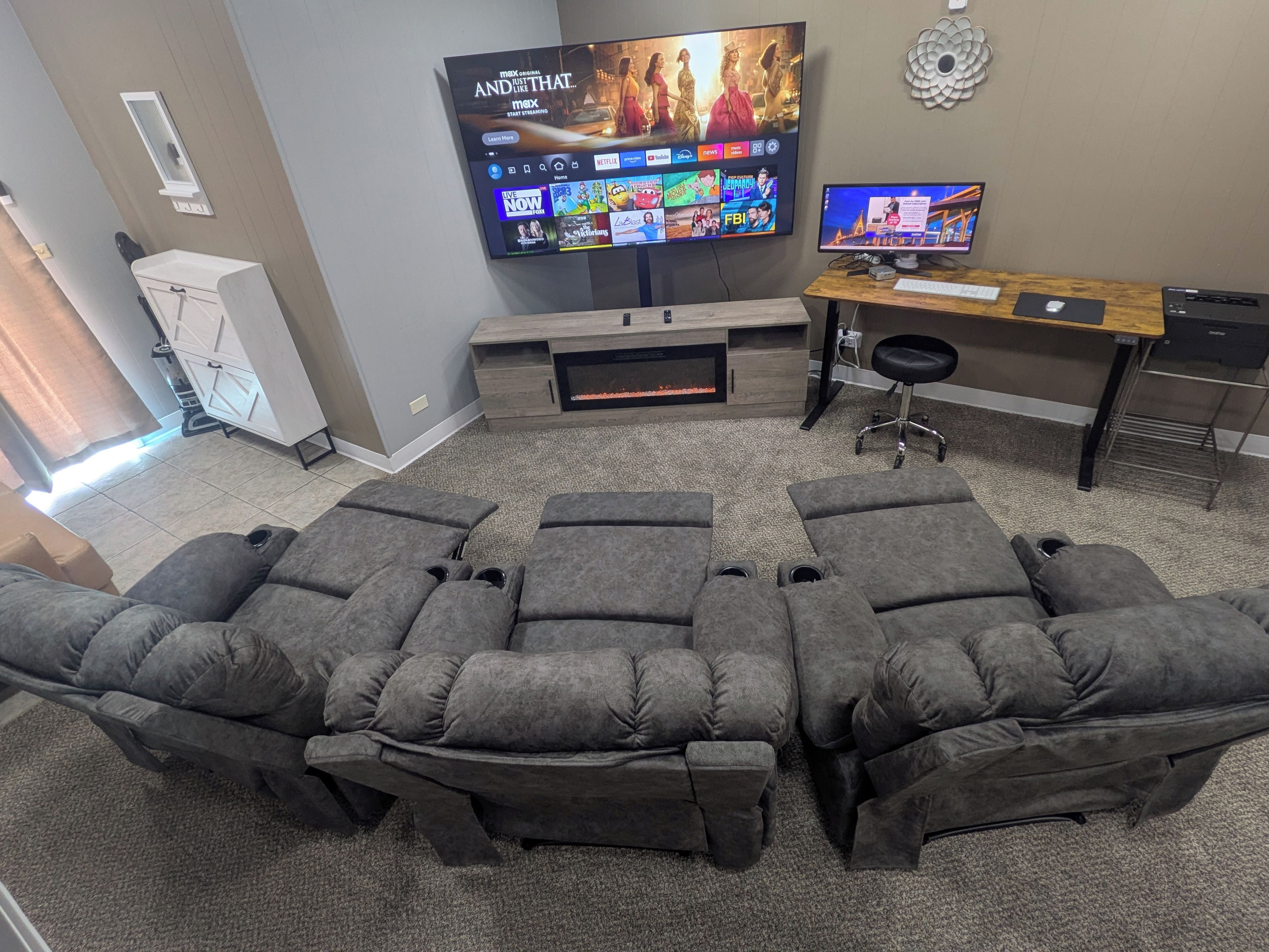 Een woonkamer met een groot flatscreen-televisiobehang aan de wand, een houten bureau met een computer, en een hoekbank.