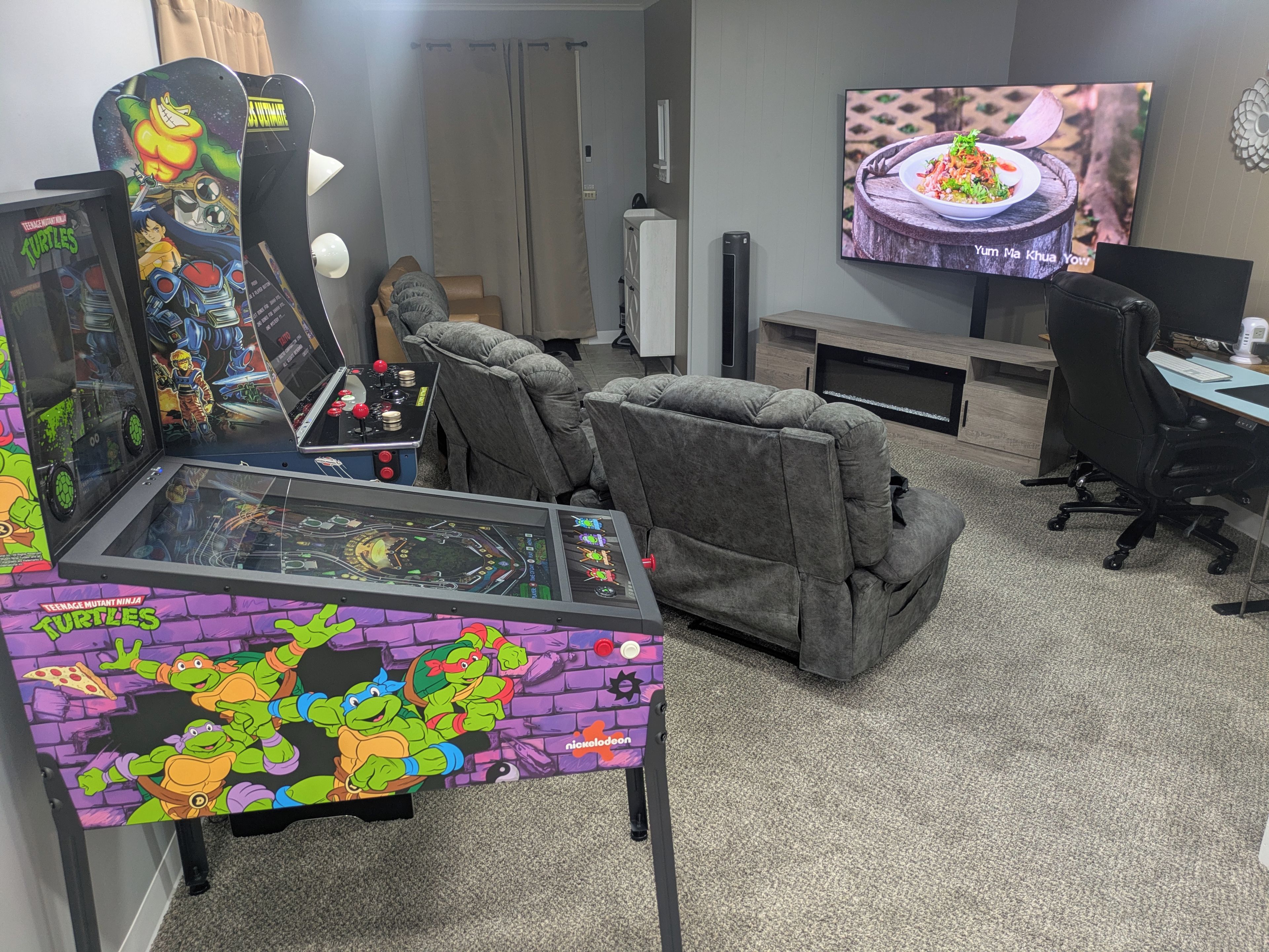 A gaming area with a racing simulator, two recliner chairs, a TV displaying a food scene, and a desk with a computer.
