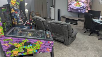 A gaming area with a racing simulator, two recliner chairs, a TV displaying a food scene, and a desk with a computer.