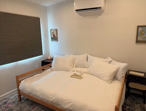 Un dormitorio con cama de madera, ropa de cama blanca, una mesita de noche, un aire acondicionado, una ventana con persianas y un cuadro en la pared.